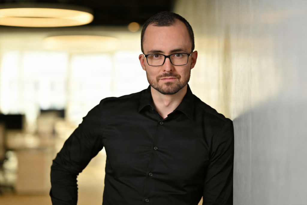 Callboy Alexander im schwarzen Hemd, an eine Bürowand gelehnt.