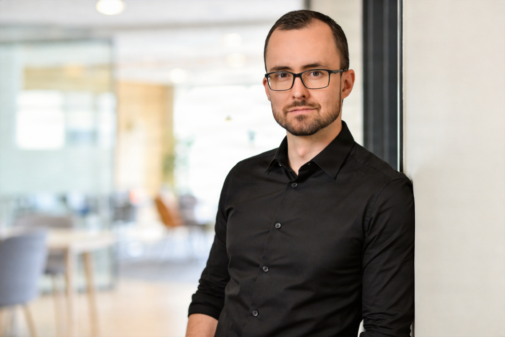 Callboy Alexander im schwarzen Hemd, leicht lächelnd an eine Bürowand gelehnt.